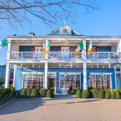 A blue house with white trim, featuring multiple flags and a landscaped front yard under a clear blue sky.