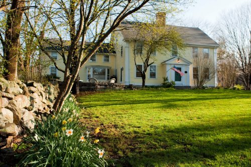 A yellow house with an American flag, surrounded by green grass and blooming flowers, under a clear blue sky.