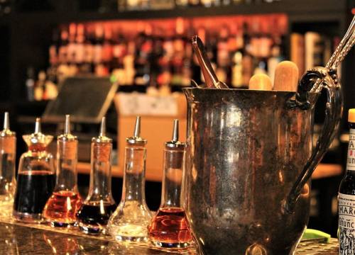 A bar setup featuring various glass bottles of ingredients, a metal shaker, and a blurred background of shelves with drinks.