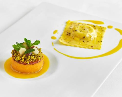 A gourmet dish featuring a round pastry topped with herbs and a ravioli, drizzled with sauce on a white plate.