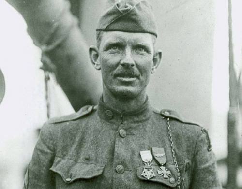 A historical black-and-white photo of a soldier in uniform, wearing medals and a cap, standing proudly.