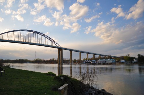 A bridge spans a river under a cloudy sky, with green grass and a building visible along the water's edge.
