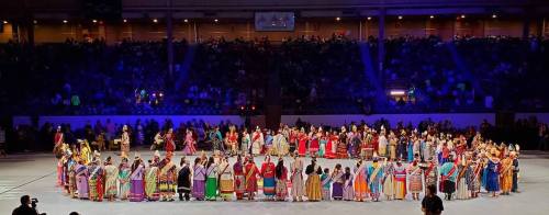 A large group of dancers in colorful traditional attire performing on a stage, with a cheering audience in the background.