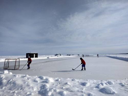 Two players in red jerseys skate on an outdoor ice rink surrounded by snow, with a goal in the foreground.