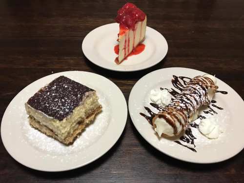 Three desserts on plates: a slice of strawberry cheesecake, a piece of tiramisu, and a chocolate-drizzled cannoli.