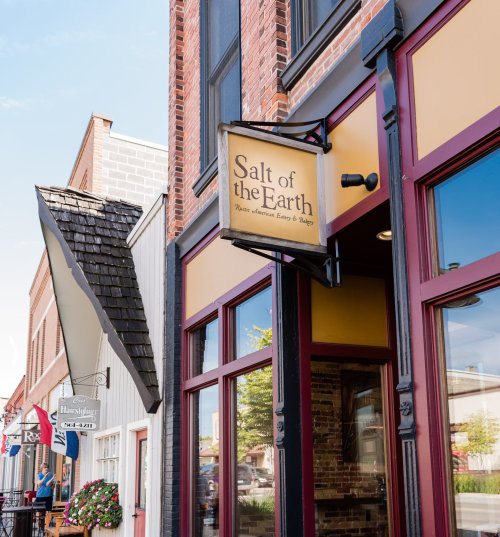Sign for "Salt of the Earth," a rustic American eatery and bakery, on a charming street with brick buildings.