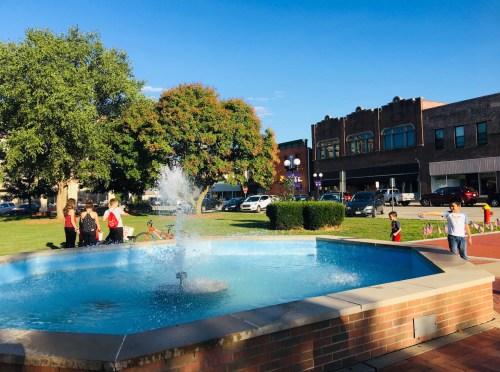 A fountain in a park with families and children playing, surrounded by trees and historic buildings under a clear blue sky.
