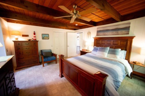 Cozy bedroom with wooden beams, a large bed, dresser, and a chair, featuring warm lighting and beach-themed decor.