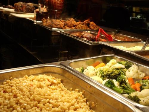 A buffet display featuring various dishes, including macaroni, mixed vegetables, and meats in metal trays.