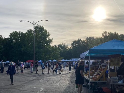 A bustling outdoor market with tents, people browsing, and a bright sun in the sky. Trees line the background.