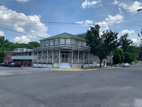 Historic building with a green roof, surrounded by trees, located at a street corner under a blue sky with clouds.
