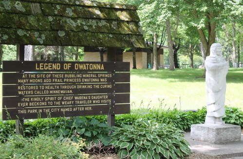 Sign about the Legend of Owatonna with a statue nearby, surrounded by greenery in a park setting.
