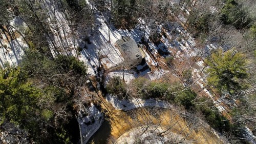 Aerial view of a snowy landscape with a small house surrounded by trees and a winding dirt path.