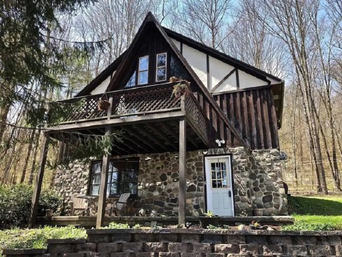 A charming two-story cabin with a stone and wood exterior, surrounded by trees and featuring a front porch.