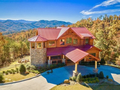 A large, rustic mountain home with a red metal roof, surrounded by trees and mountains under a clear blue sky.