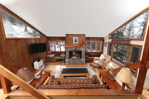 Cozy cabin interior with a stone fireplace, wooden walls, and large windows overlooking a snowy landscape.