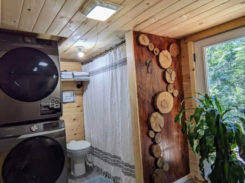 A cozy bathroom with a shower curtain, wooden wall decor, laundry machines, and a plant by the window.