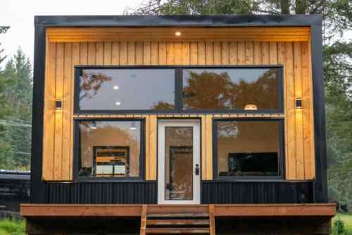 Modern cabin with large windows, wooden siding, and a welcoming entrance, surrounded by trees.
