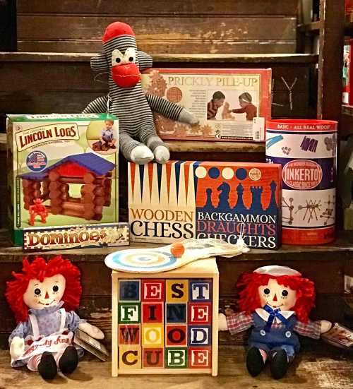 A collection of classic toys including Lincoln Logs, a sock monkey, and wooden games, displayed on a wooden shelf.