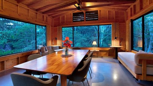A cozy wooden interior with large windows, a dining table, and a sofa, surrounded by lush greenery outside.