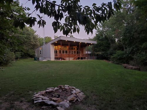 A modern house with a porch, surrounded by trees, and a stone fire pit in the foreground on a grassy lawn.