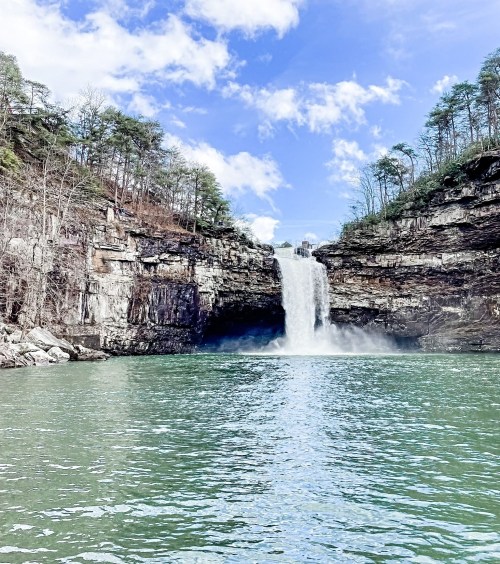 A stunning waterfall cascades into a serene turquoise lake, surrounded by rocky cliffs and lush trees under a blue sky.