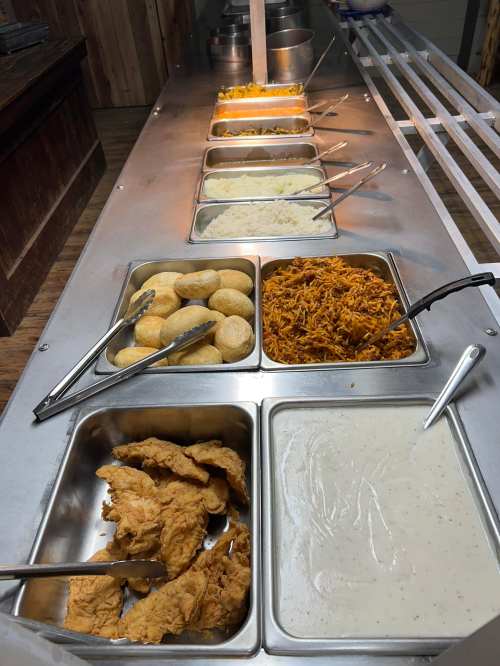 A buffet spread featuring rice, chicken, shredded meat, rolls, and sauces in metal trays with serving utensils.