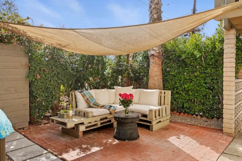 Cozy outdoor seating area with a pallet sofa, table, and a bouquet of roses under a shade sail. Lush greenery surrounds.