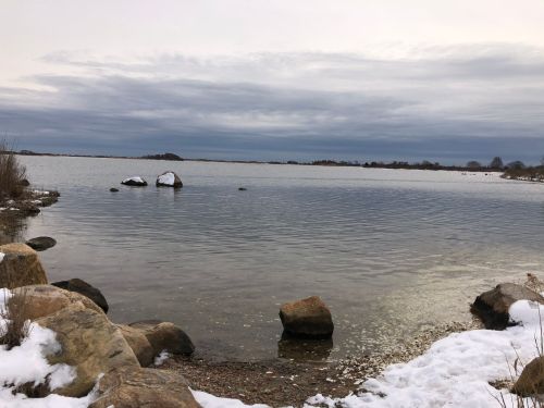 A calm lake scene with rocky shores and patches of snow under a cloudy sky.