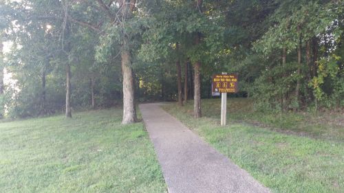 A paved path leads into a wooded area, with a sign indicating park rules and directions.