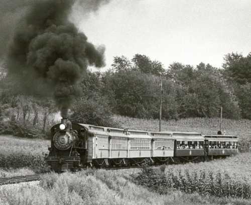 A vintage steam train billows black smoke as it travels through a rural landscape with trees and fields.