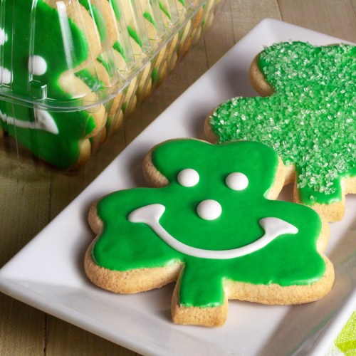 Green shamrock-shaped cookies with smiley faces, decorated with icing and sugar, on a white plate.
