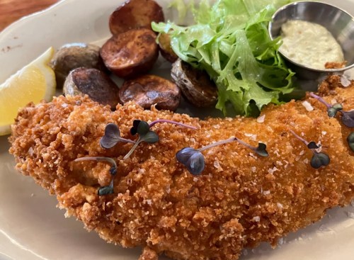 A plate featuring crispy fried fish, roasted potatoes, fresh greens, a lemon wedge, and a small cup of dipping sauce.