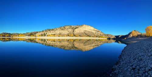 A serene lake reflecting a rocky hill under a clear blue sky, surrounded by autumn foliage.