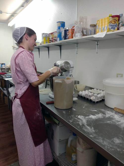 A woman in a pink dress and apron mixes ingredients in a kitchen, surrounded by baking supplies and eggs.