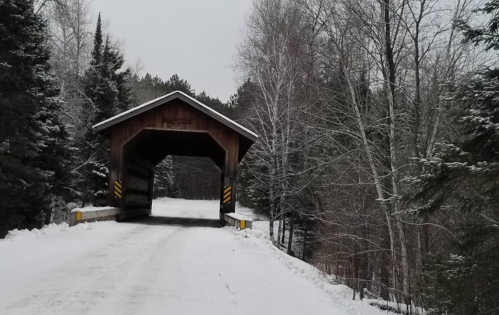 A snow-covered road leads to a wooden covered bridge surrounded by trees in a winter landscape.
