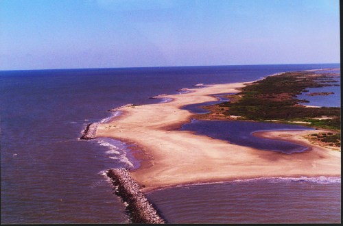 Aerial view of a sandy peninsula extending into the ocean, surrounded by water and greenery.
