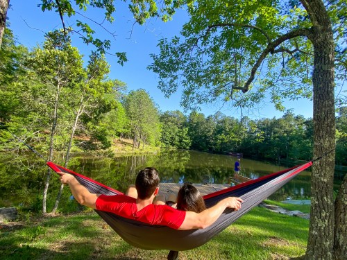 A couple relaxes in a hammock by a serene pond, surrounded by lush trees and a clear blue sky.