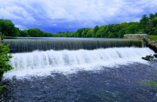 A serene waterfall cascades over a dam, surrounded by lush green trees and a cloudy blue sky.