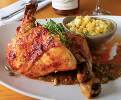 A plate featuring a roasted chicken leg with sauce, garnished with greens, alongside a bowl of corn and creamy dip.