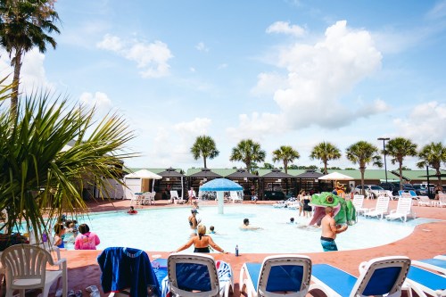 A lively pool scene with people swimming and lounging on chairs, surrounded by palm trees and sunny skies.