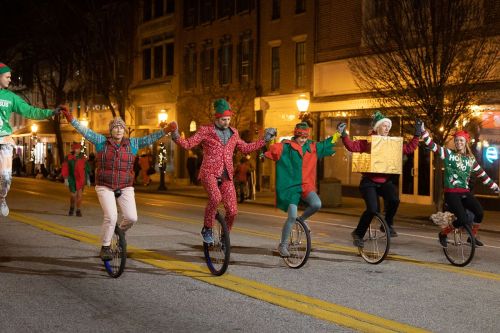 A festive group of performers in holiday attire riding unicycles and holding hands during a nighttime parade.