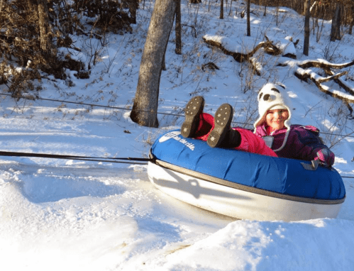 A child in a panda hat enjoys tubing down a snowy hill, smiling as they glide on a blue inflatable tube.