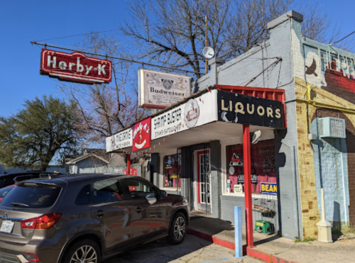 A small storefront with signs for "Herby-K," "Liquors," and "Grill & Burger," alongside a parked car and clear blue sky.