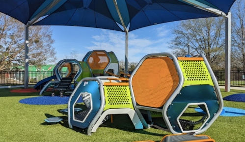 Colorful playground equipment under a large blue canopy, set on green turf with a clear blue sky in the background.