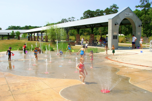 Children play in a splash pad, running through water jets on a sunny day, with adults supervising nearby.
