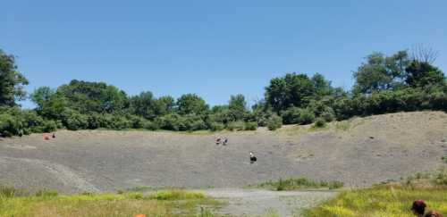 A large, rocky crater surrounded by greenery, with a few people exploring the area. Bright blue sky overhead.