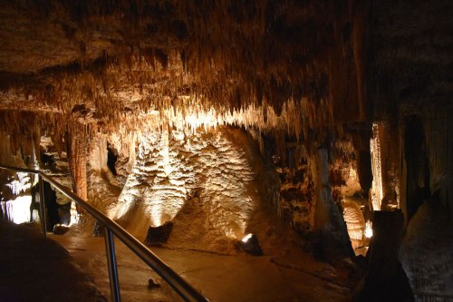 A dimly lit cave interior with stalactites and stalagmites, featuring a railing along a pathway.