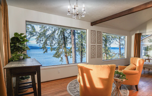 Cozy living space with orange chairs, large windows overlooking a lake, and trees, featuring warm lighting and decor.