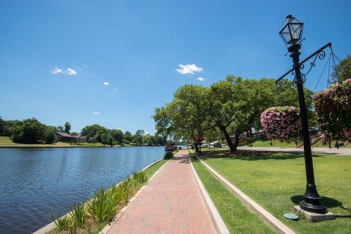 A scenic riverside path with a lamp post, greenery, and hanging flower baskets under a clear blue sky.